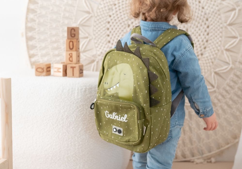 Kind trägt grünen Dino Kindergartenrucksack mit Namen Gabriel, Rückansicht im Kinderzimmer